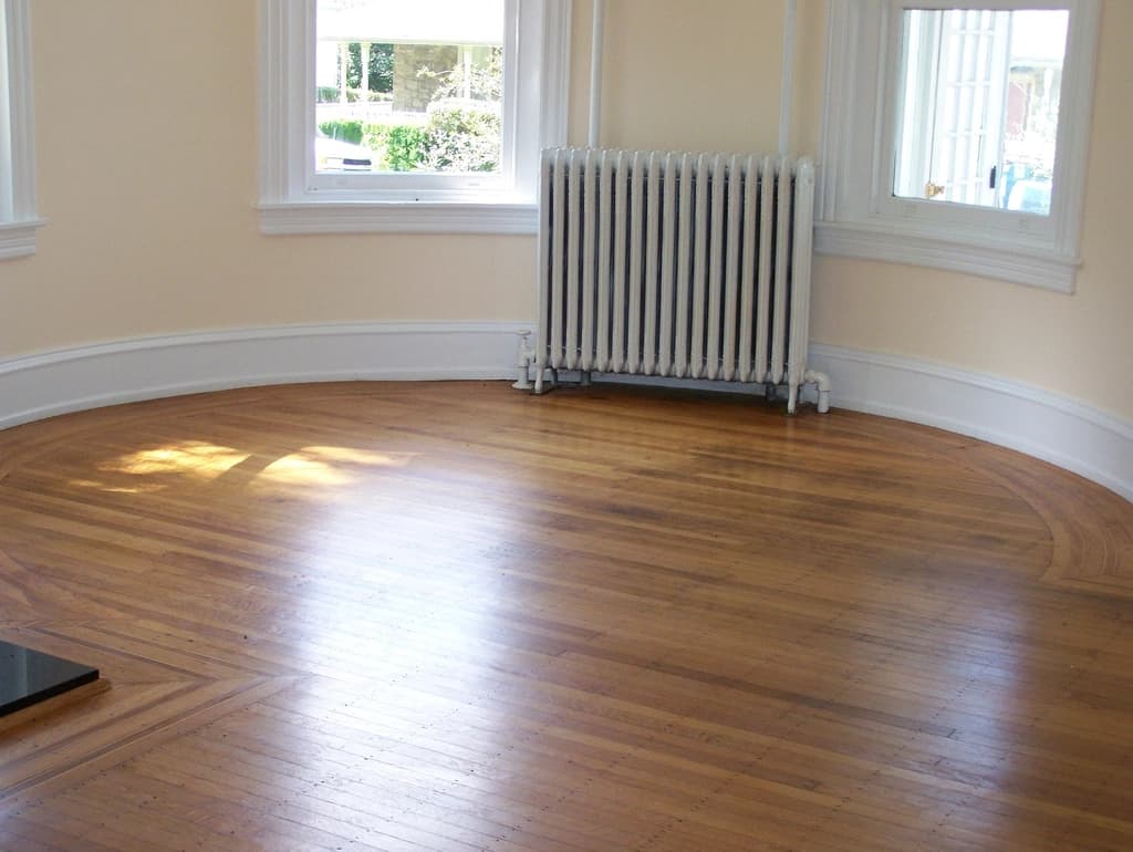 A bright, empty living room with a bay window, a radiator and a wooden floor