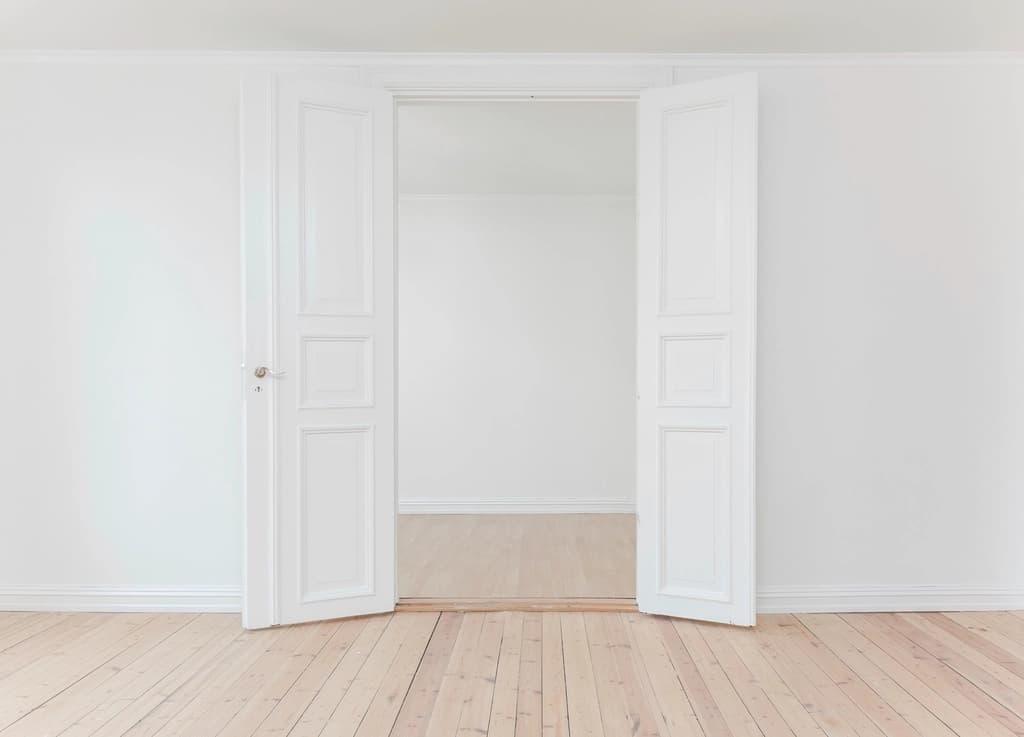 An empty white room with double doors open onto another empty room